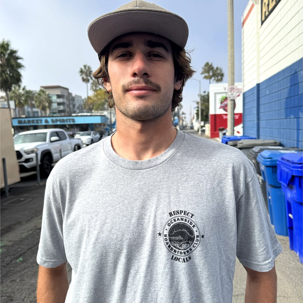 Man wearing a gray t-shirt with a logo and text, standing outdoors with palm trees and buildings in the background.