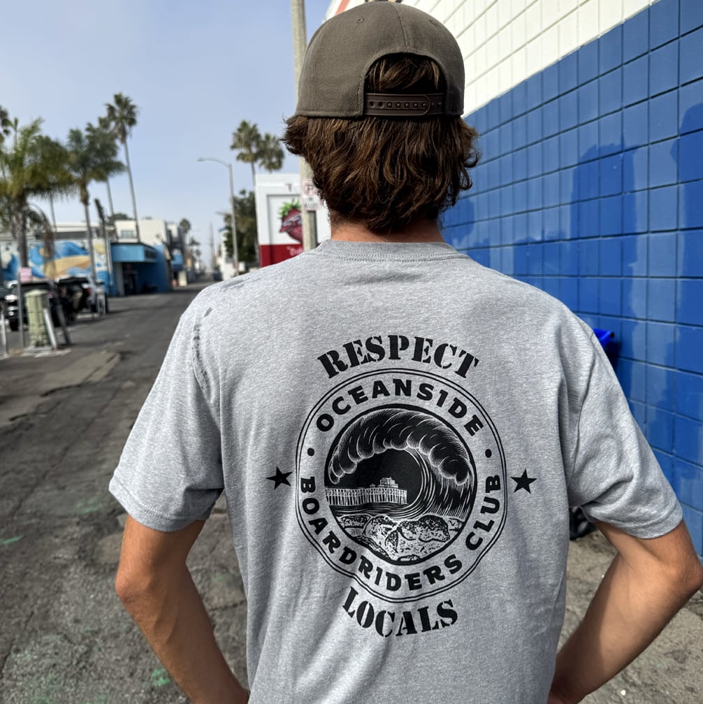 Person wearing a gray t-shirt with 'Respect Oceanside Boardriders Club Locals' design outdoors.